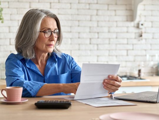 An older woman sits at the kitchen counter looking at a piece of paper. A laptop, a mug, and a calculator are on the counter.