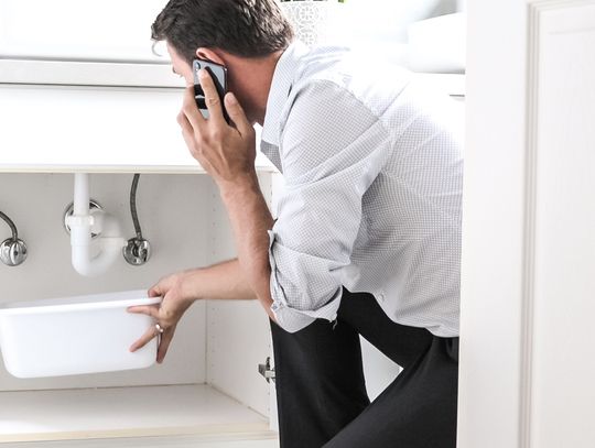 A homeowner kneeling under his bathroom sink, holding a bucket to catch water. He's talking on the phone with the other hand.