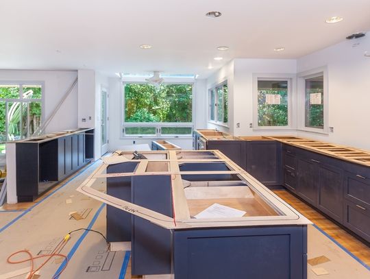 A kitchen that’s under renovation; all the counters are missing from the cabinets, and cardboard covers the floor.