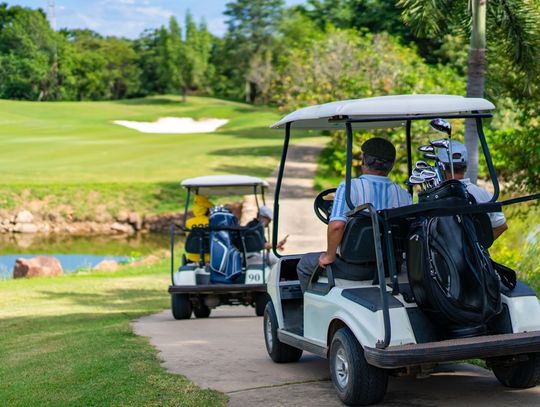 A group of four people in two separate golf carts is riding through a golf course cart path near a faux lake.