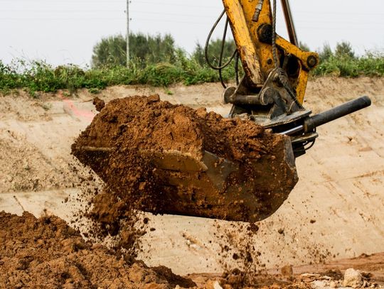 A yellow excavator bucket scoops clay-rich soil at a construction site with dirt mounds and vegetation in the background.