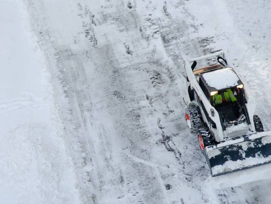 An overhead view of a compact tractor with a front-mounted snow plow operating in frigid, snowy winter conditions.