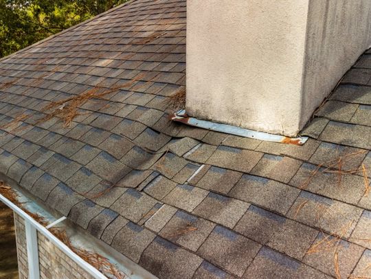A weathered shingle roof with loose chimney flashing, scattered pine needles, and debris collected in the gutter.