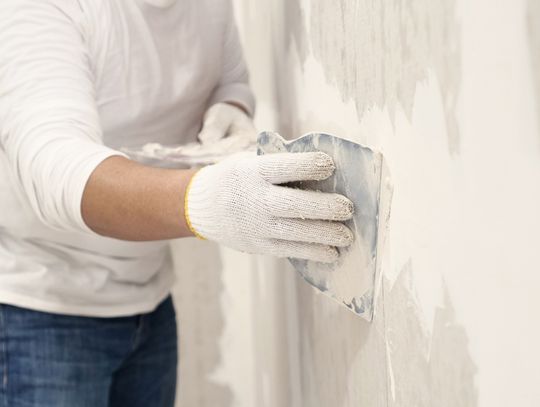 Person wearing gloves applies joint compound to a drywall surface with a taping knife while smoothing wall repairs indoors.