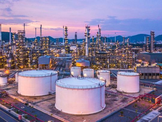 An aerial view of a large oil and gas refinery at twilight. The refinery has several chemical tanks and vertical pipes.