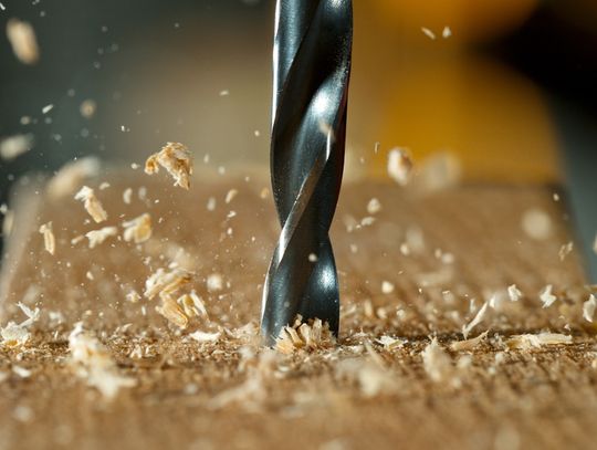 Close-up of a sharp drill bit boring into a wooden board as wood shavings and sawdust scatter through the air.
