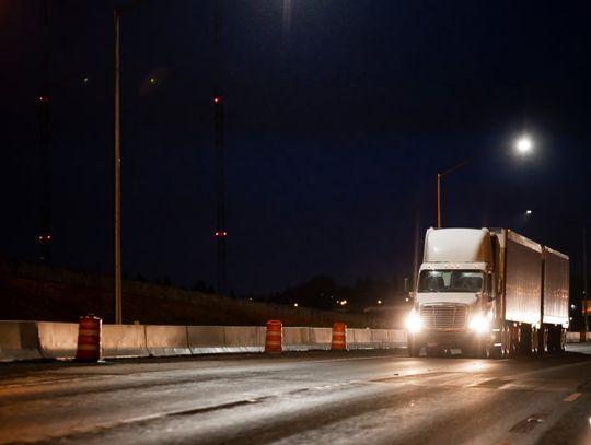 A white semi-truck with its headlights on driving next to road barricades and orange and white traffic barrels at night.