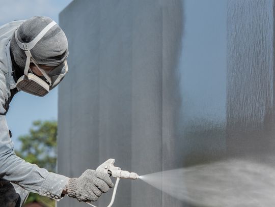 Worker wearing protective gear spray-coats a large metal surface with an industrial coating outdoors.