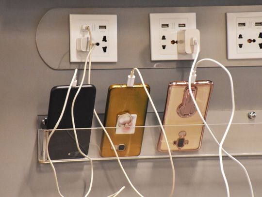 Three smartphones connected to charging cables at a public charging station with multiple electrical outlets.