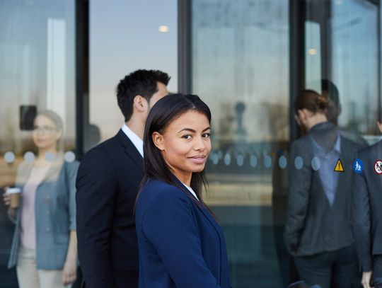 A woman dressed in business professional attire looks to the side while walking through an automatic door.