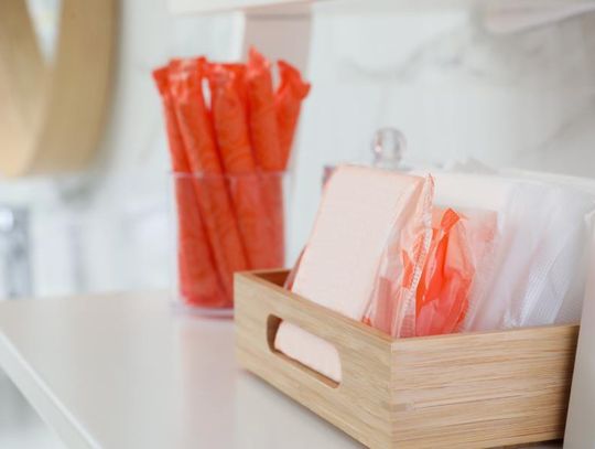 A small wooden crate contains menstrual pads of various sizes. A glass jar contains tampons in orange wrappers.