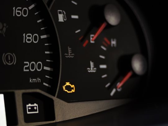 A close-up focos on a vehicle dashboard. A small yellow check engine light displays beside the speedometer.