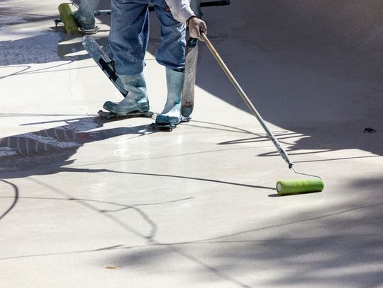 A worker in rubber boots uses a long-handled roller to apply coating to an empty concrete swimming pool surface.