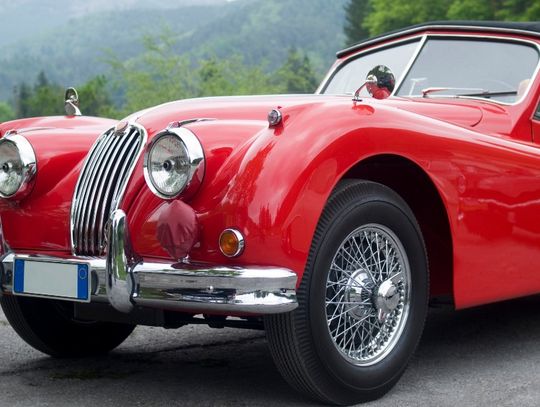 A red classic convertible car with chrome details and wire-spoke wheels parked near a wooded mountain area.