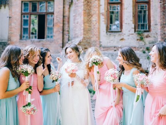 A bride stands outside beside her six bridesmaids; three bridesmaids wear blue dress and three wear pink.
