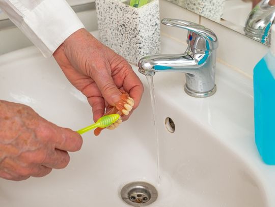 An individual is using a yellow toothbrush to brush their dentures. They are holding their dentures over a sink.