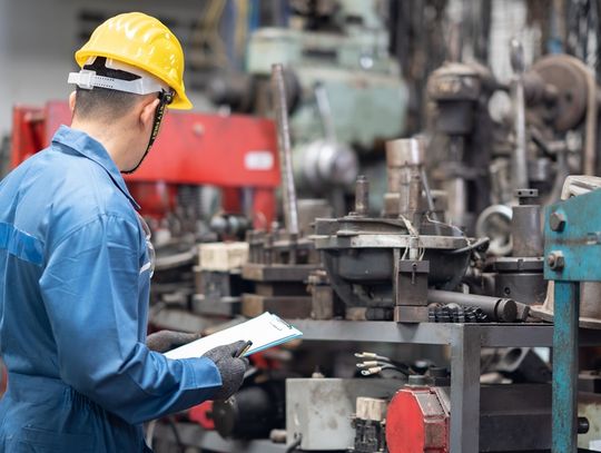 A person wearing a yellow hard hat is holding a small clipboard and inspecting metal machine components.