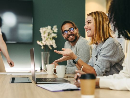 Coworkers gather around a conference table with laptops, coffee cups, and papers. One person gestures while speaking.