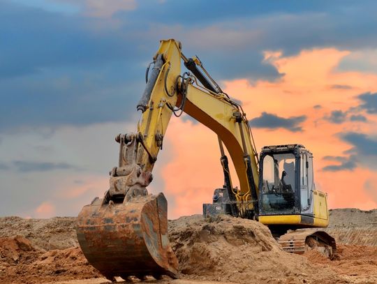 A yellow excavator digging into large mounds of brown soil beneath a cloudy sky with orange light near the horizon.