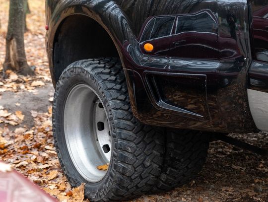A close-up of the back tires of a pickup truck with dually wheels parked in a forest over dry leaves next to trees.
