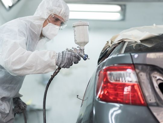 A person in protective gear spraying a gray car body with a paint gun inside a bright automotive paint booth.