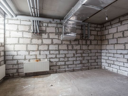 An unfinished basement with exposed concrete block walls, overhead pipes, HVAC ductwork, radiators, and a bare concrete floor.