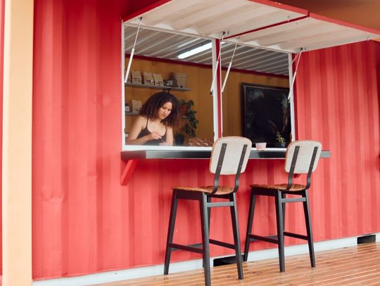 Woman working inside a red shipping container restaurant with an open service window and two stools outside.