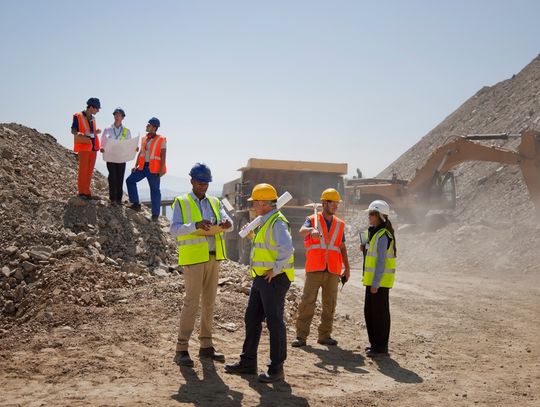 Several construction workers in hard hats and safety vests reviewing plans at a rocky excavation site with heavy machinery.