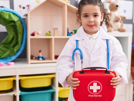 A little girl dressed up as a doctor and holding a red first aid kit in front of her as she stands in a classroom.