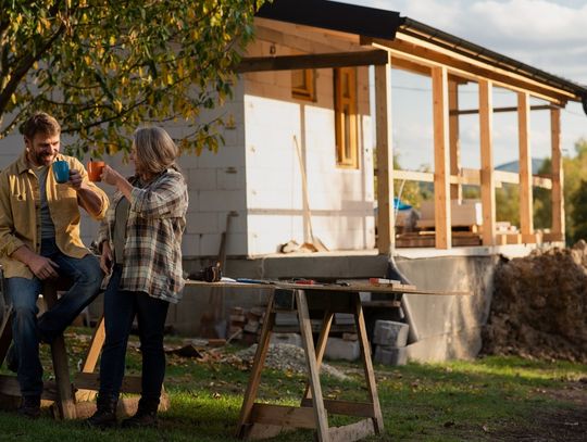 People holding mugs near a work table and trees. There is an unfinished house with wood posts behind the couple.