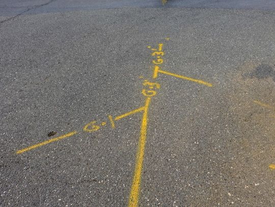 A patch of concrete with yellow spray paint indicating utility lines nearby. The lines also have numbers and letters.
