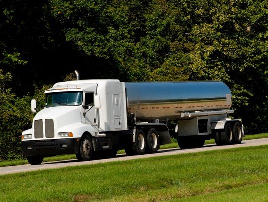 A tanker trailer with a white cab and silver fuel tank drives along a road between a treeline and grassy divider.