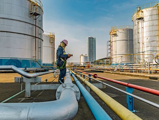 a worker in safety gear using a tablet among colorful pipelines and large storage tanks at an industrial site.