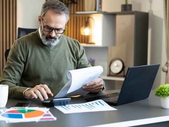 A business owner reviews financial paperwork with a calculator and laptop while charts sit across his office desk.