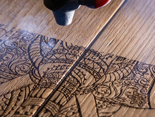 A black and red laser engraving machine is engraving an intricate pattern into a piece of light-colored wood.