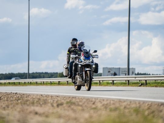 A motorcycle with a small windshield carrying the rider and a passenger in the back down a sunny concrete road.
