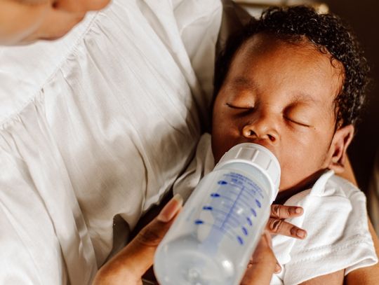 A parent cradles a sleeping baby in her arms. The baby still has an empty milk bottle in their mouth.