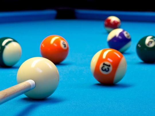 A close-up of a Billiard pool table, and someone is aiming their pool stick towards the orange, striped 13-ball.