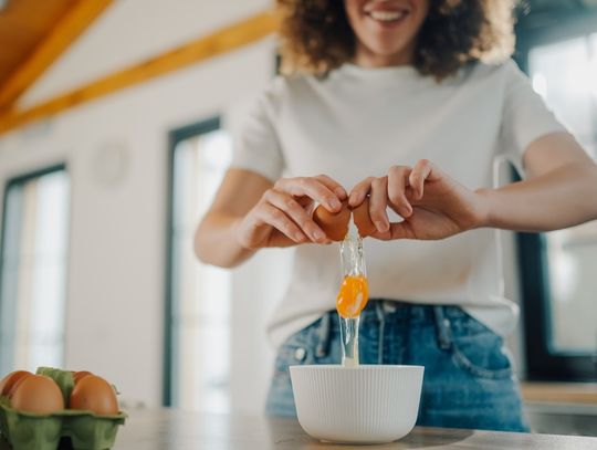 A woman wearing a white shirt cracks a brown egg into a bowl on a kitchen counter beside an egg carton.