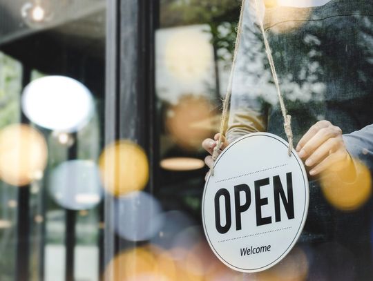 A hand turning an open sign on a glass door, with a person in an apron and blurred cafe interior behind it.