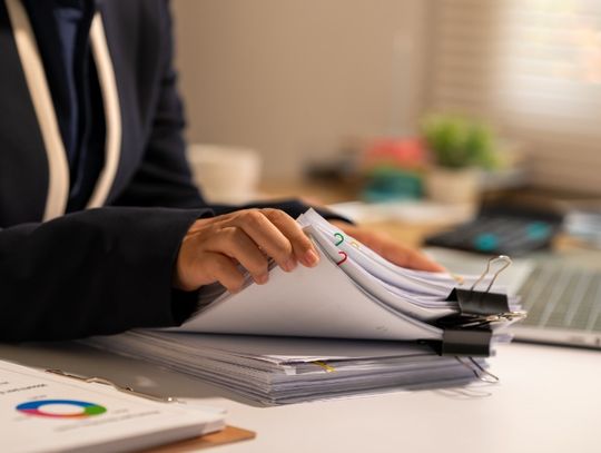 A woman sits at a desk, flipping through papers. There is a laptop blurry in the background.