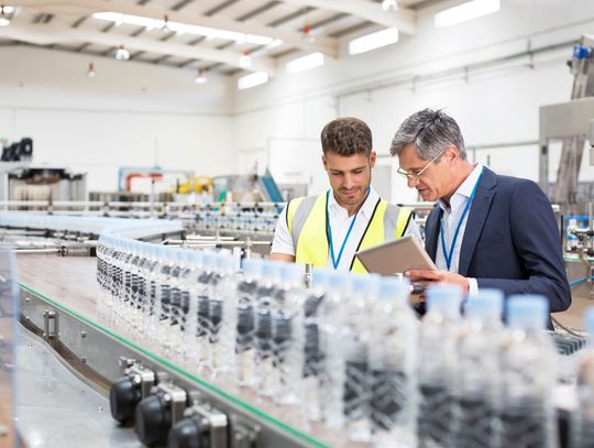 Two men stand behind a manufacturing line with hundreds of plastic water bottles. One person holds a tablet.