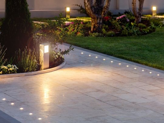 A residential walkway is illuminated by small, circular lights that are in the pavers. Other lighting appears in the landscaping.