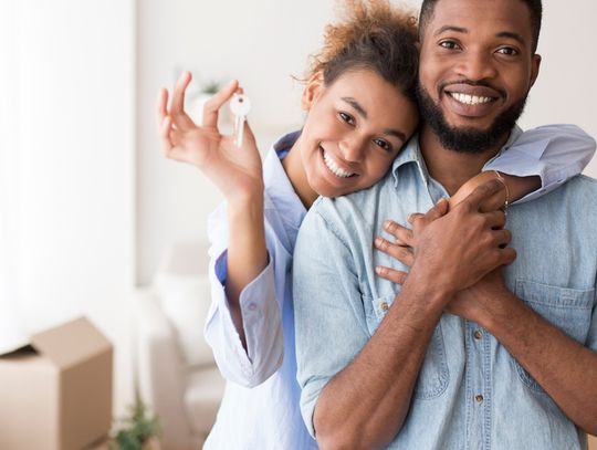 A smiling woman stands behind a smiling man and has one arm draped around his shoulders and the other holding up a key.