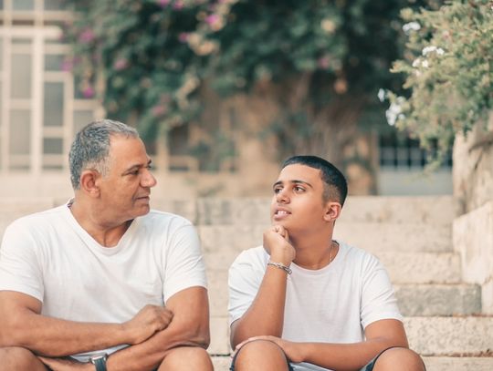 A teenage son looking thoughtfully toward his aging father as they sit on the stone steps of a home and converse.