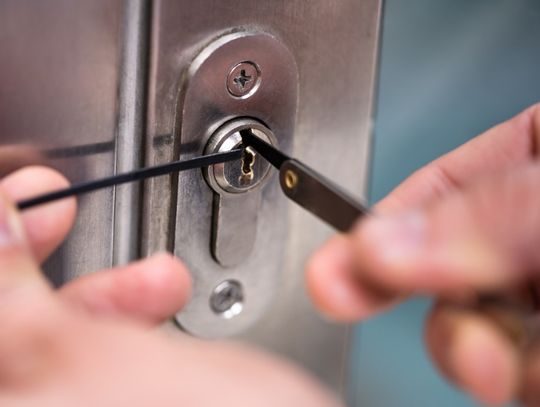 A close-up of two hands holding slender metal tools inside the keyhole of a silver door lock mounted on a metal door.