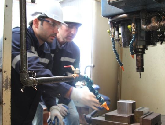 Two Caucasian men wearing hard hats and safety goggles observe a large machine surrounded by metal walls.