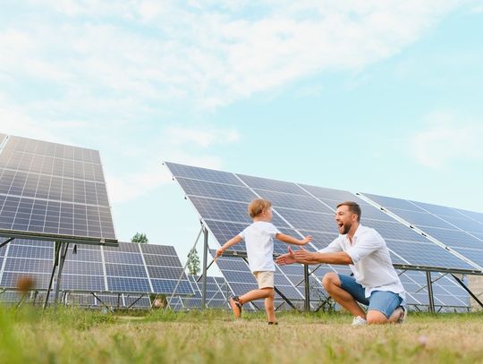 A man is kneeling down as a small child runs into his open arms. They are outside, in front of a set of solar panels.