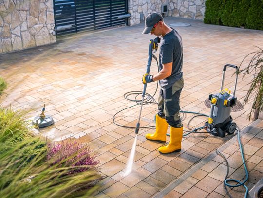 A man wearing yellow rubber boots pressure washes a brick patio beside landscaping, with cleaning equipment nearby.