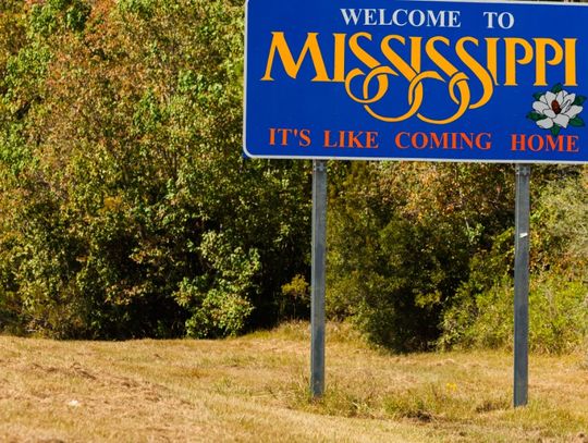 A big blue road sign state sign along the highway. The sign reads "Welcome to Mississippi, it's like coming home."